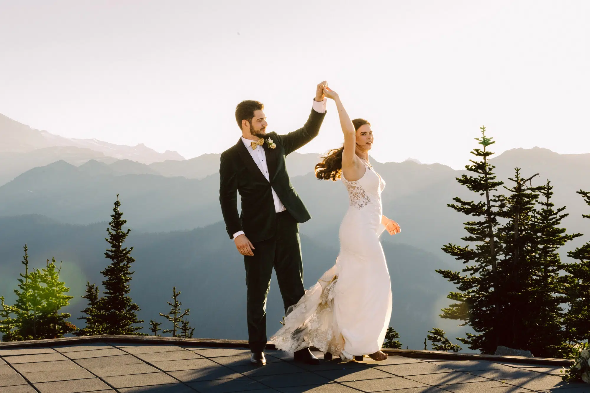 Snohomish Wedding Photographer GSquared Weddings A bride in a stunning wedding dress and a groom in a dark suit dance outdoors on a mountain ledge at sunset, surrounded by evergreen trees and scenic peaks. Serving Seattle, Snohomish and Orlando