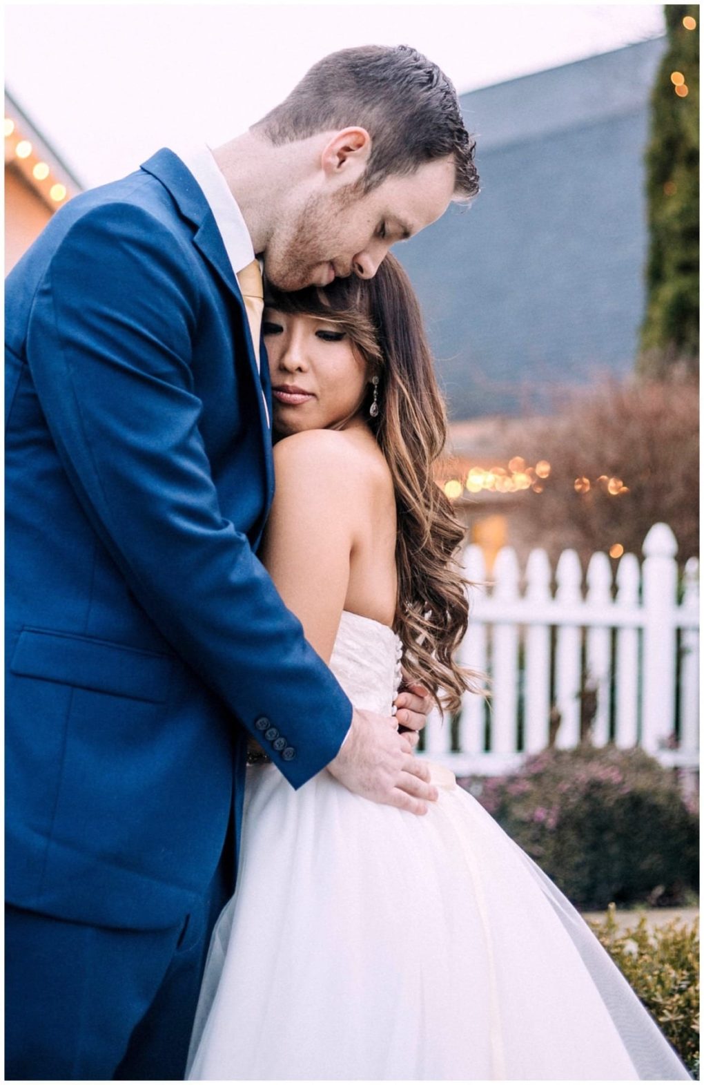 Premier snohomish & seattle wedding photographer | gsquared weddings photography a groom in a blue suit gently embraces and kisses the forehead of his bride in a white dress as they stand outside near a white picket fence surrounded by soft lights and greenery at their snohomish seattle wedding | serving snohomish seattle                   & beyond