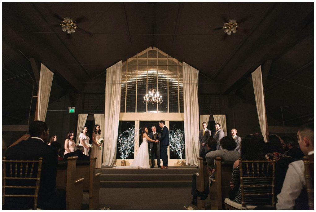 Premier snohomish & seattle wedding photographer | gsquared weddings photography a bride and groom stand at the altar holding hands during their snohomish wedding ceremony surrounded by bridesmaids and groomsmen in a warmly lit indoor venue with draped curtains and a chandelier overhead Guests are seated in the pews | serving snohomish seattle                   & beyond