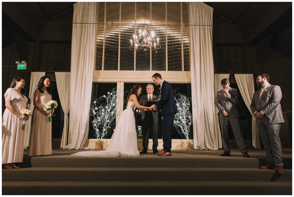 Premier snohomish & seattle wedding photographer | gsquared weddings photography a bride and groom stand facing each other holding hands on an indoor stage with a chandelier white drapes and string lights surrounded by bridesmaids and groomsmen during their romantic snohomish wedding ceremony near seattle | serving snohomish seattle                   & beyond