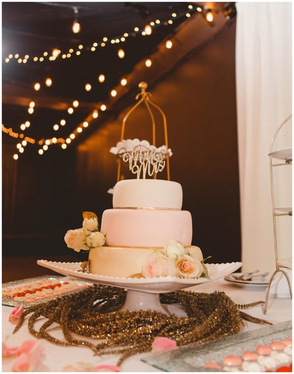 Premier snohomish & seattle wedding photographer | gsquared weddings photography a three tiered white wedding cake with pastel pink accents and roses topped with a gold mr & mrs sign sits on a decorated table under string lights at a warmly lit snohomish seattle wedding reception | serving snohomish seattle                   & beyond