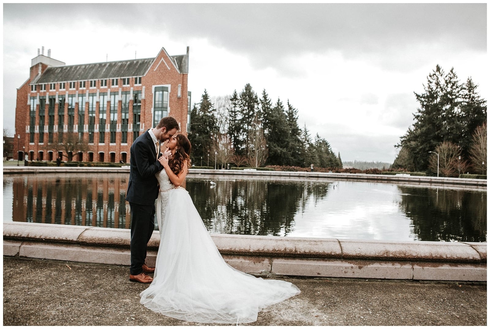 Premier Snohomish & Seattle Wedding Photographer | GSquared Weddings Photography A bride and groom stand close together, embracing beside a reflective pond at a Snohomish Seattle wedding, with a large brick building and trees in the background under a cloudy sky.                                   | Serving Snohomish, Seattle ,                  & Beyond