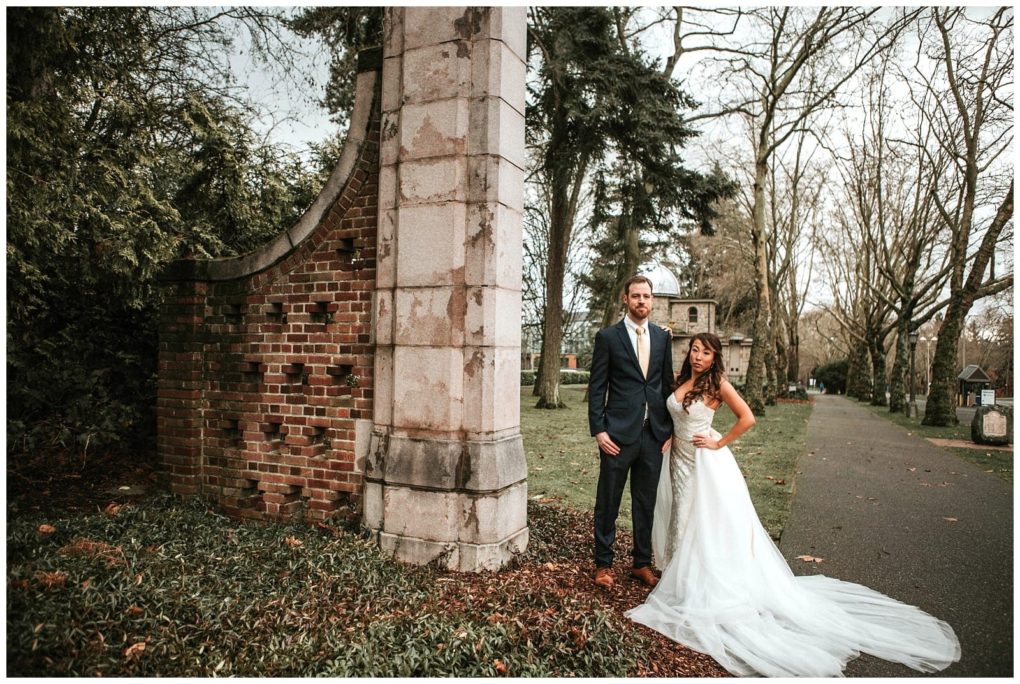 Premier snohomish & seattle wedding photographer | gsquared weddings photography a bride in a white gown and groom in a suit stand together near a tall brick and stone structure in a park with trees and a paved path on a cloudy day capturing their snohomish seattle wedding engagement | serving snohomish seattle                   & beyond