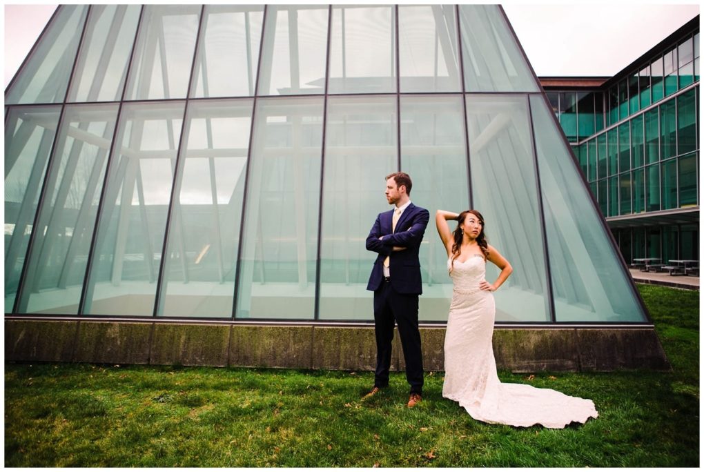 Premier snohomish & seattle wedding photographer | gsquared weddings photography a bride in a white gown stands with one hand on her hip beside a groom in a suit both posing confidently on a grassy lawn The modern glass building behind them sets the scene for their stylish snohomish seattle wedding engagement | serving snohomish seattle                   & beyond