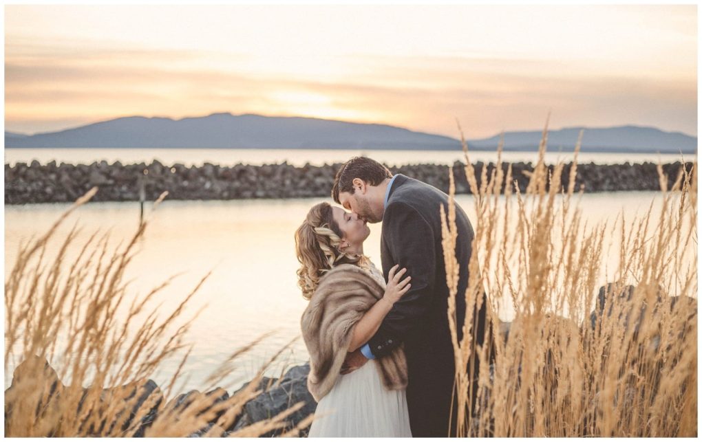 Premier snohomish & seattle wedding photographer | gsquared weddings photography a bride and groom embrace and kiss by a lake at sunset framed by tall golden grasses with mountains and a stone breakwater in the backgroundcapturing the romance of a snohomish seattle wedding engagement moment | serving snohomish seattle                   & beyond