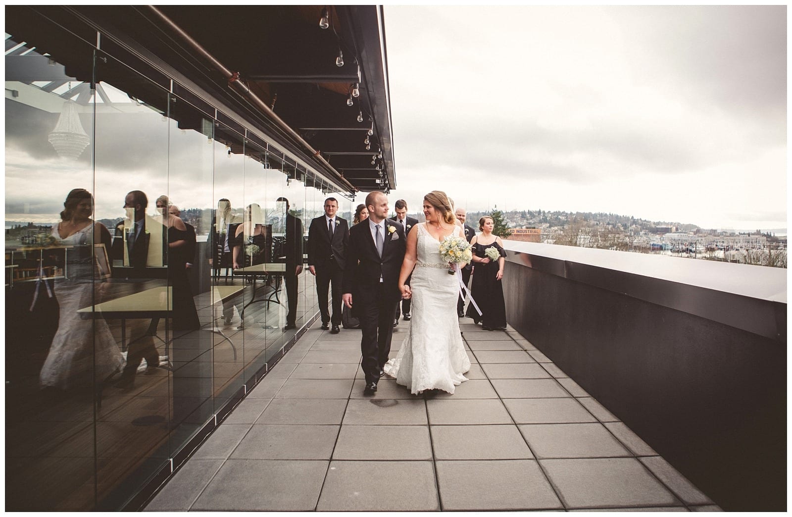 Premier Snohomish & Seattle Wedding Photographer | GSquared Weddings Photography A bride and groom walk together on a rooftop terrace in Snohomish, smiling and holding hands, while bridesmaids and groomsmen follow behind. The scene is beautifully reflected in the glass wall beside them under a cloudy Seattle sky.                                   | Serving Snohomish, Seattle ,                  & Beyond
