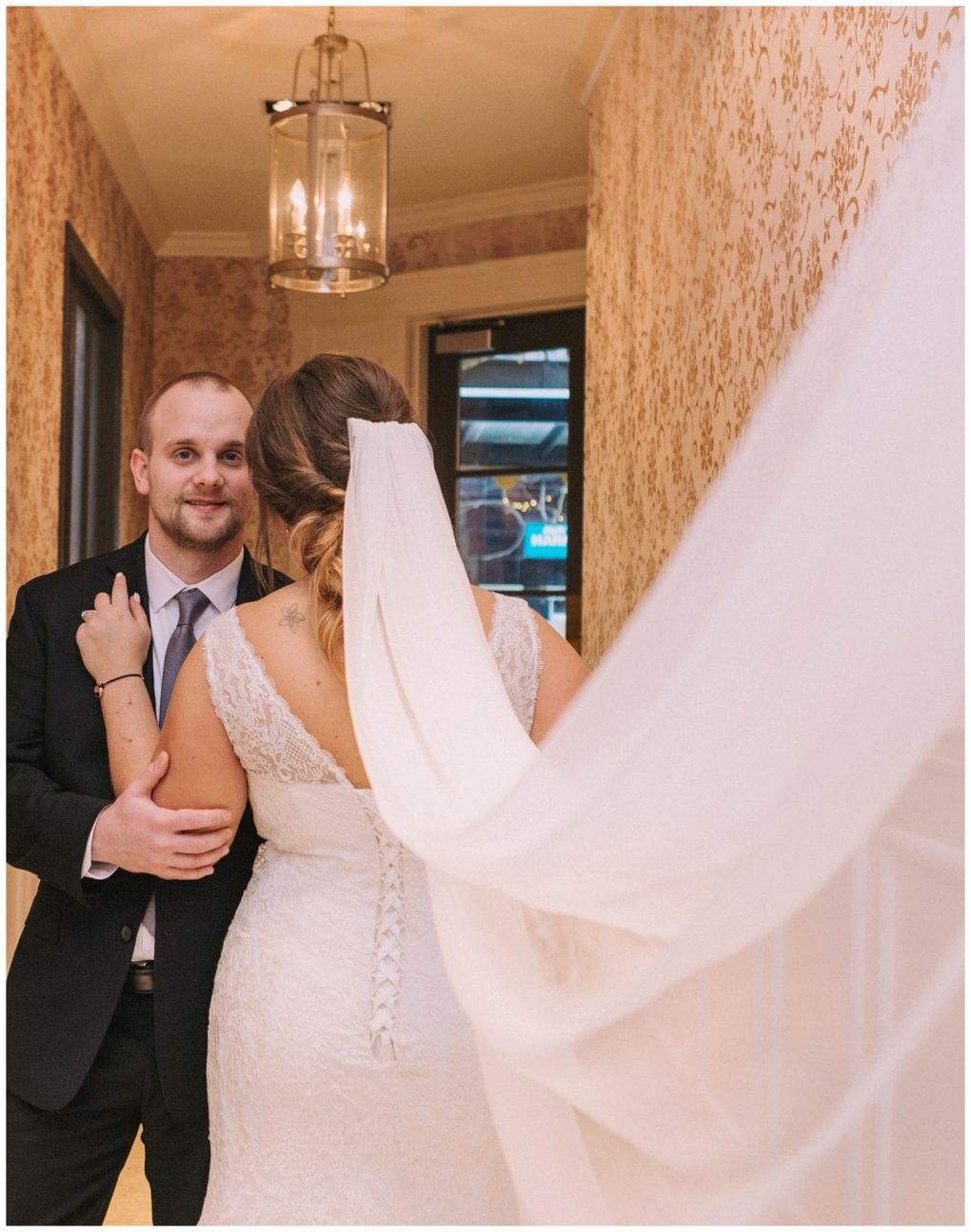 Premier snohomish & seattle wedding photographer | gsquared weddings photography a groom in a black suit smiles while holding his bride who faces away in a lace wedding dress with a long veil flowing behind her They stand in a hallway with patterned wallpapera romantic snohomish seattle wedding moment | serving snohomish seattle                   & beyond
