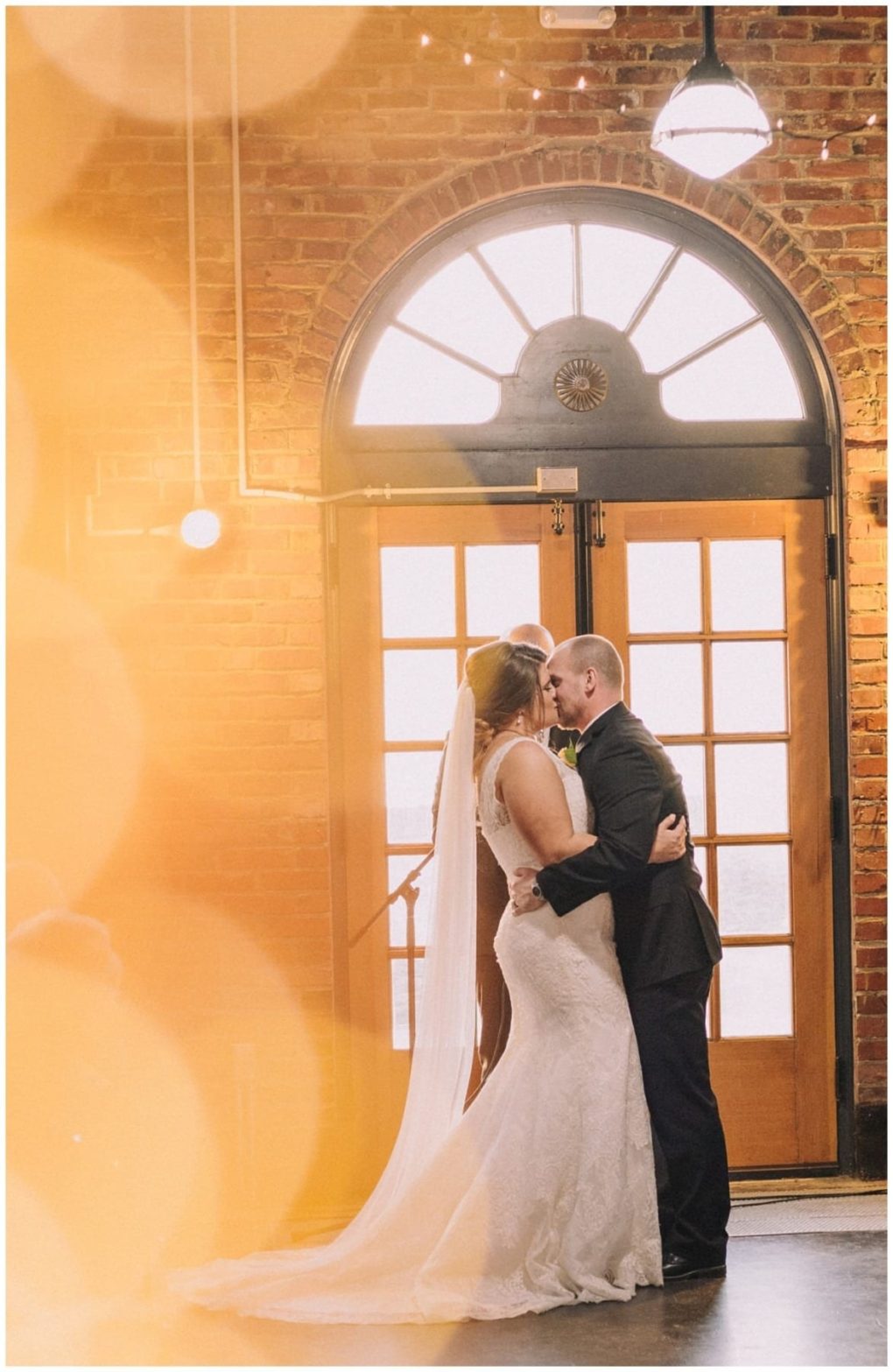 Premier snohomish & seattle wedding photographer | gsquared weddings photography a bride and groom embrace and gaze at each other during their snohomish wedding in front of a sunlit arched window with warm golden bokeh lights in the foreground and exposed brick walls behind them | serving snohomish seattle                   & beyond