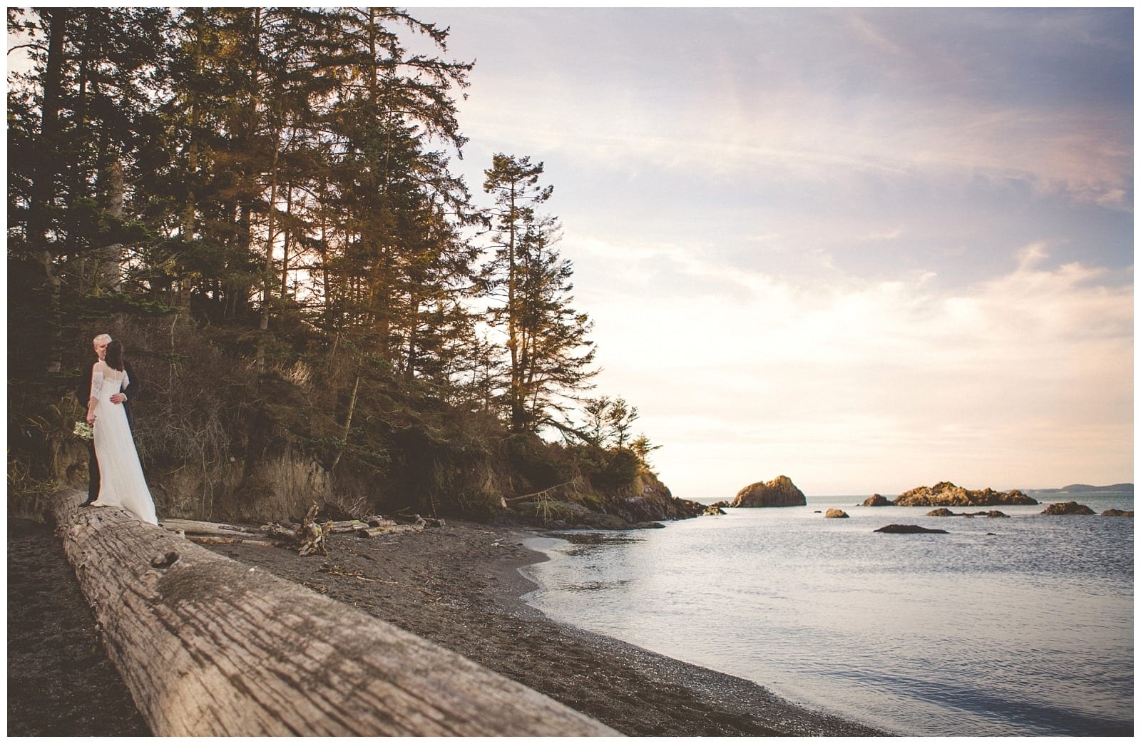 Premier Snohomish & Seattle Wedding Photographer | GSquared Weddings Photography A bride in a white dress stands on a quiet Snohomish beach near tall trees, holding a bouquet. The sky is soft and pastel as the sun sets over the calm ocean and distant rocks—perfect for a Seattle wedding engagement.                                   | Serving Snohomish, Seattle ,                  & Beyond