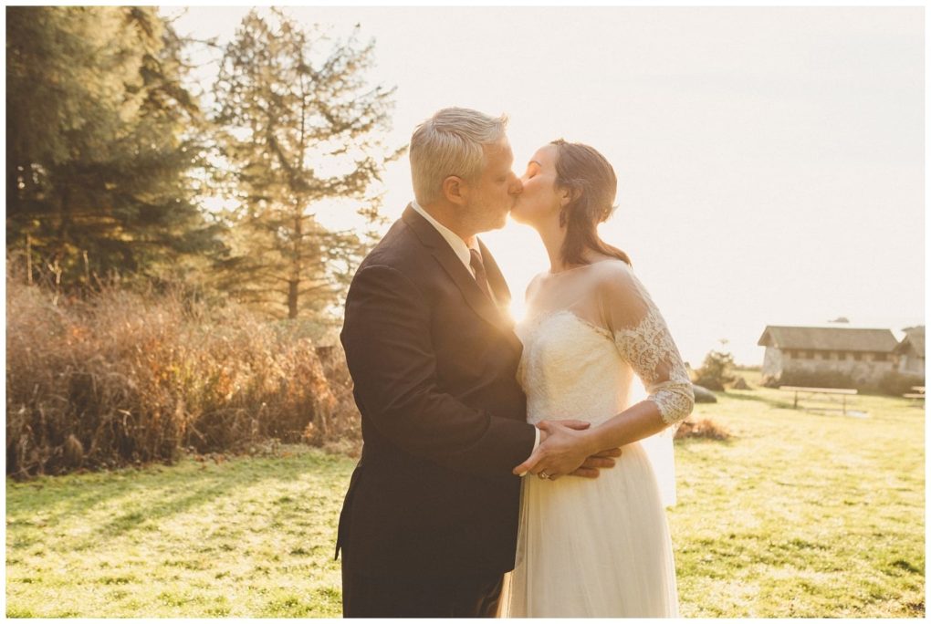 Premier snohomish & seattle wedding photographer | gsquared weddings photography a bride and groom kiss outdoors in warm sunlight at their snohomish wedding surrounded by trees and grass The bride wears a white dress with lace sleeves and the groom is in a dark suit The scene has a soft romantic and natural atmosphere | serving snohomish seattle                   & beyond