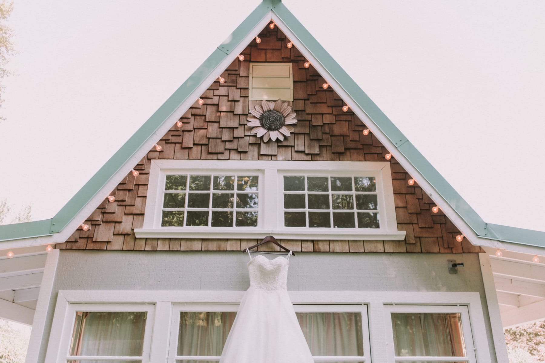 A white wedding dress hangs on the front of a cozy Snohomish house with a triangular roof, decorative shingles, string lights, and a sunflower ornament above the windows. Snohomish Wedding Photography
