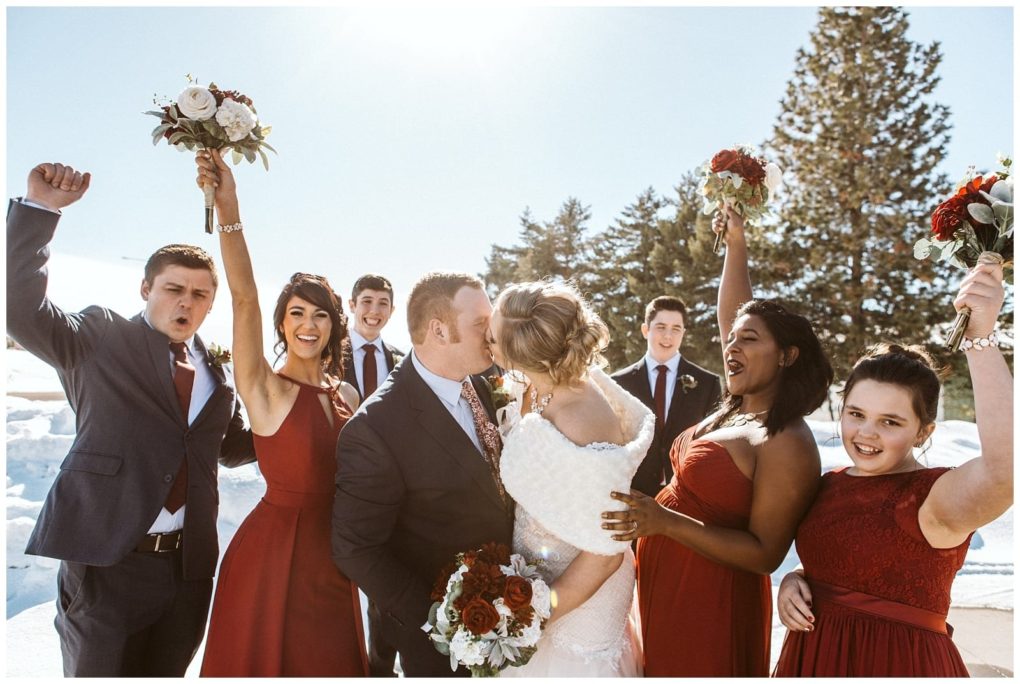 Premier snohomish & seattle wedding photographer | gsquared weddings photography a bride and groom kiss outdoors in the snow at their snohomish wedding surrounded by a joyful party in red dresses and dark suits raising bouquets and cheering under a bright sunny seattle sky | serving snohomish seattle                   & beyond
