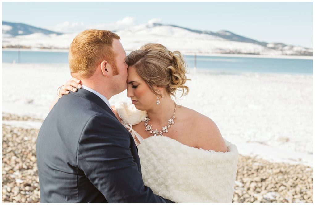 Premier snohomish & seattle wedding photographer | gsquared weddings photography a groom kisses the brides forehead as they embrace outdoors in winter near snohomish with snow covered ground and mountains behind them The bride wears a white shawl and floral necklace both appear emotional and serene during their seattle wedding | serving snohomish seattle                   & beyond