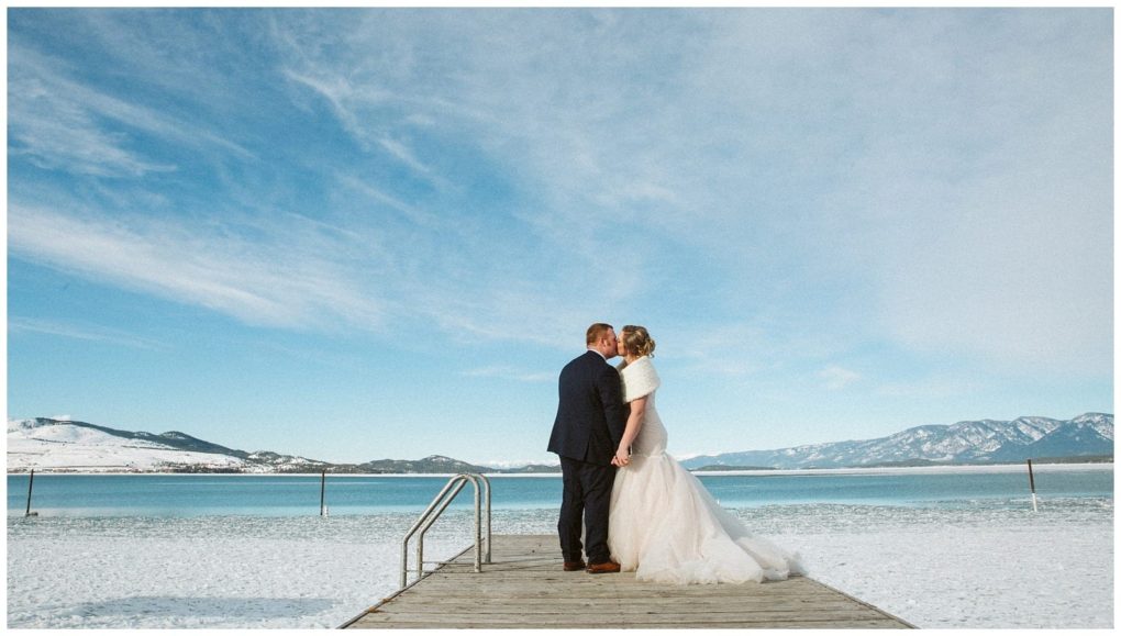 Premier snohomish & seattle wedding photographer | gsquared weddings photography a bride and groom stand on a snowy dock by a lake holding hands and kissing Snow covered mountains and a bright blue sky create a serene winter landscape perfect for a magical snohomish seattle wedding moment | serving snohomish seattle                   & beyond