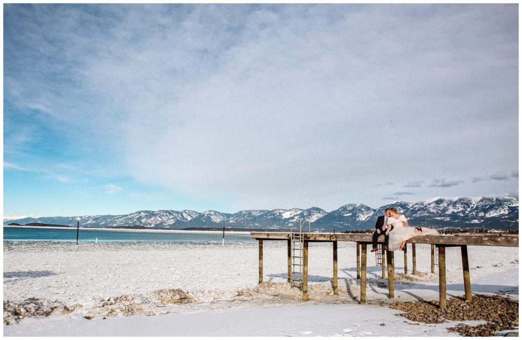 Premier snohomish & seattle wedding photographer | gsquared weddings photography a bride and groom sit together on a wooden pier over snowy ground by a lake with mountains and a bright blue sky in the backgroundcapturing the magic of a snohomish seattle wedding engagement | serving snohomish seattle                   & beyond
