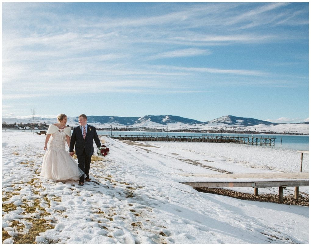 Premier snohomish & seattle wedding photographer | gsquared weddings photography a bride and groom walk hand in hand on a snowy lakeshore near snohomish with mountains and a wooden pier in the background under a blue sky with wispy cloudsa perfect seattle wedding engagement moment | serving snohomish seattle                   & beyond