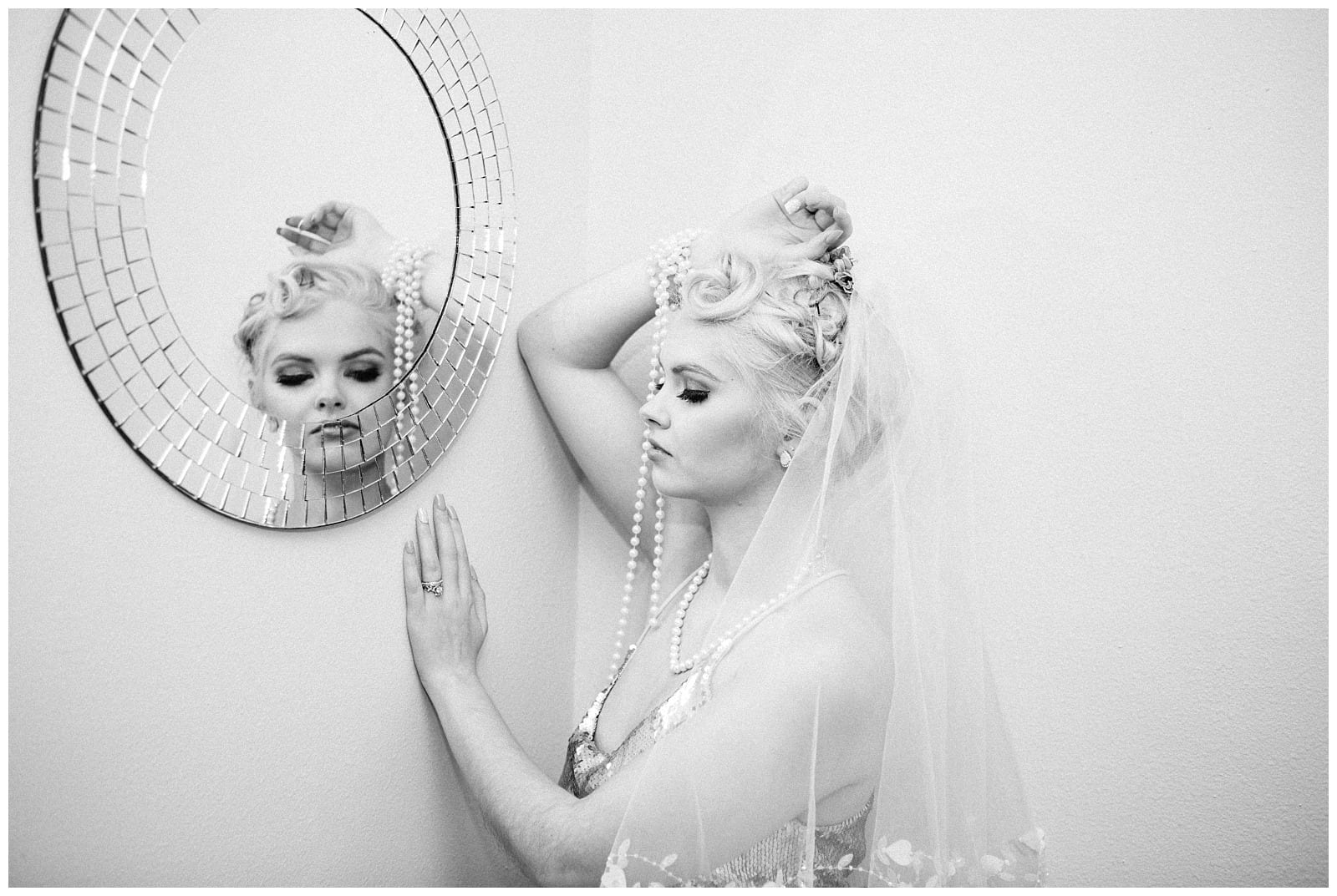 Premier Snohomish & Seattle Wedding Photographer | GSquared Weddings Photography A bride in a veil and pearls stands by a wall, gently touching a round mosaic mirror, her reflection visible as she gazes downward with a thoughtful expression. This black and white moment captures timeless Snohomish Seattle wedding elegance.                                   | Serving Snohomish, Seattle ,                  & Beyond