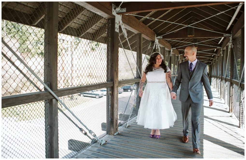 Premier snohomish & seattle wedding photographer | gsquared weddings photography a bride and groom walk hand in hand across a covered wooden bridge in snohomish smiling at each other The bride wears a white dress and purple shoes the groom wears a gray suit and tie Sunlight filters through the sides of the bridge | serving snohomish seattle                   & beyond