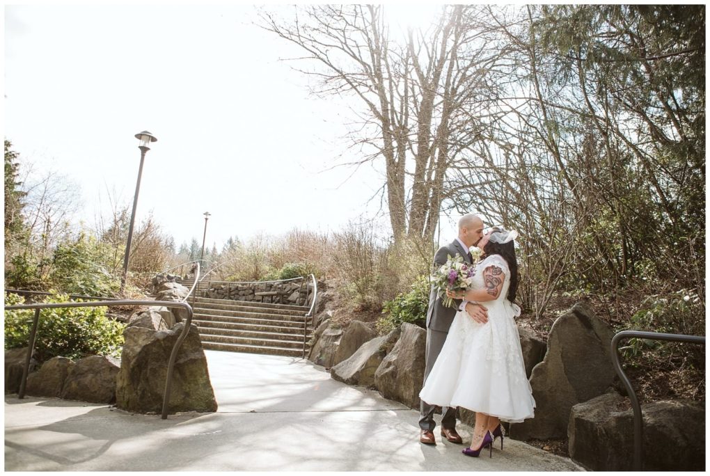 Premier snohomish & seattle wedding photographer | gsquared weddings photography a bride and groom kiss outdoors on a sunny day near stone steps surrounded by trees and bushes during their snohomish wedding The bride holds a bouquet and wears a short white dress the groom wears a vest and tie Sunlight shines above them | serving snohomish seattle                   & beyond
