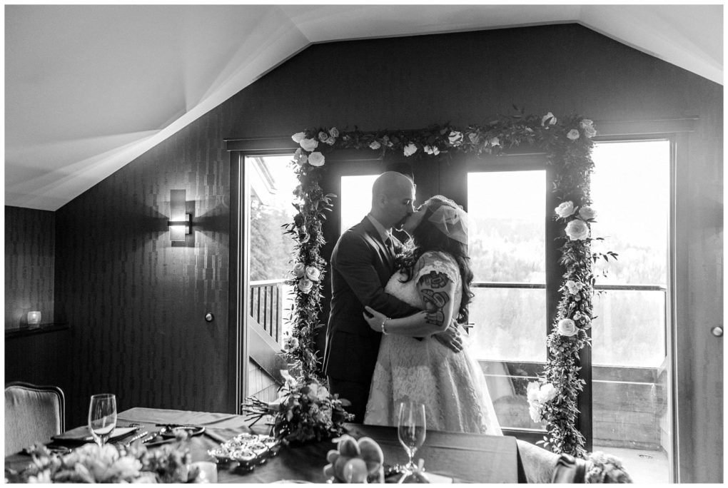 Premier snohomish & seattle wedding photographer | gsquared weddings photography a black and white photo of a snohomish bride and groom kissing in front of a floral arch by a window with a scenic view outside The bride wears a veil and lace dress the groom is in a suit A dining table is in the foreground | serving snohomish seattle                   & beyond