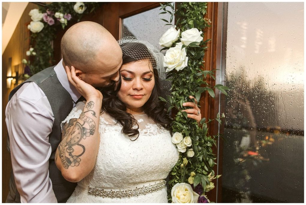 Premier snohomish & seattle wedding photographer | gsquared weddings photography a groom with tattoos gently kisses the brides cheek as she smiles softly They stand beside a floral arch and rain speckled window dressed in wedding attire at their romantic snohomish seattle wedding | serving snohomish seattle                   & beyond