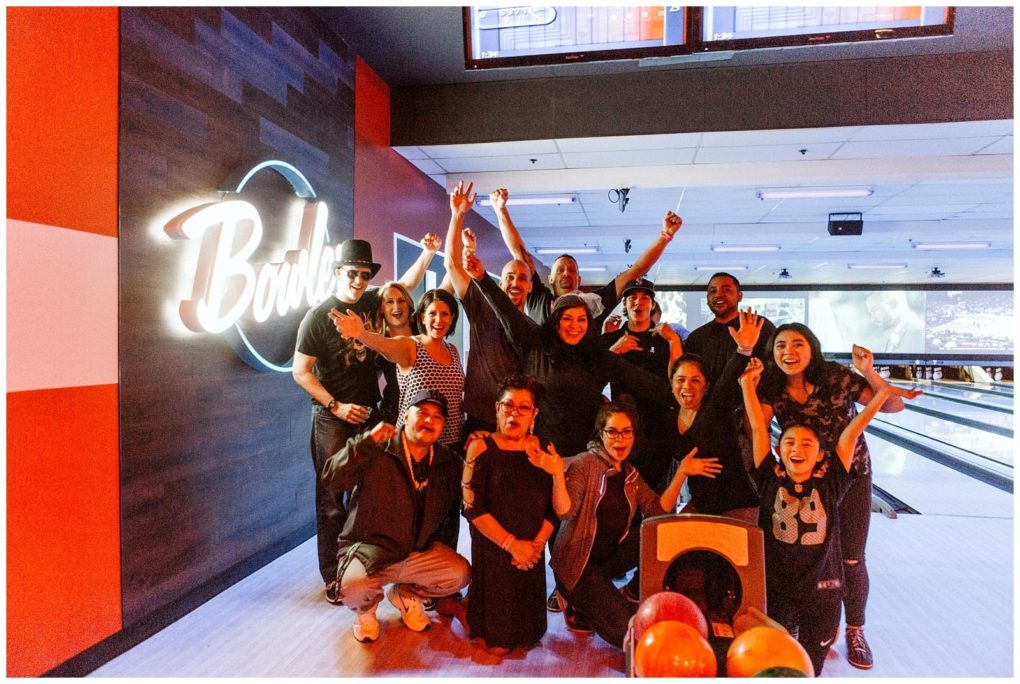 Premier snohomish & seattle wedding photographer | gsquared weddings photography a group of people posing and cheering together inside a bowling alley near snohomish with colorful bowling balls in the foreground and a large illuminated bowl sign on the wall behind themperfect for a seattle wedding or engagement celebration | serving snohomish seattle                   & beyond