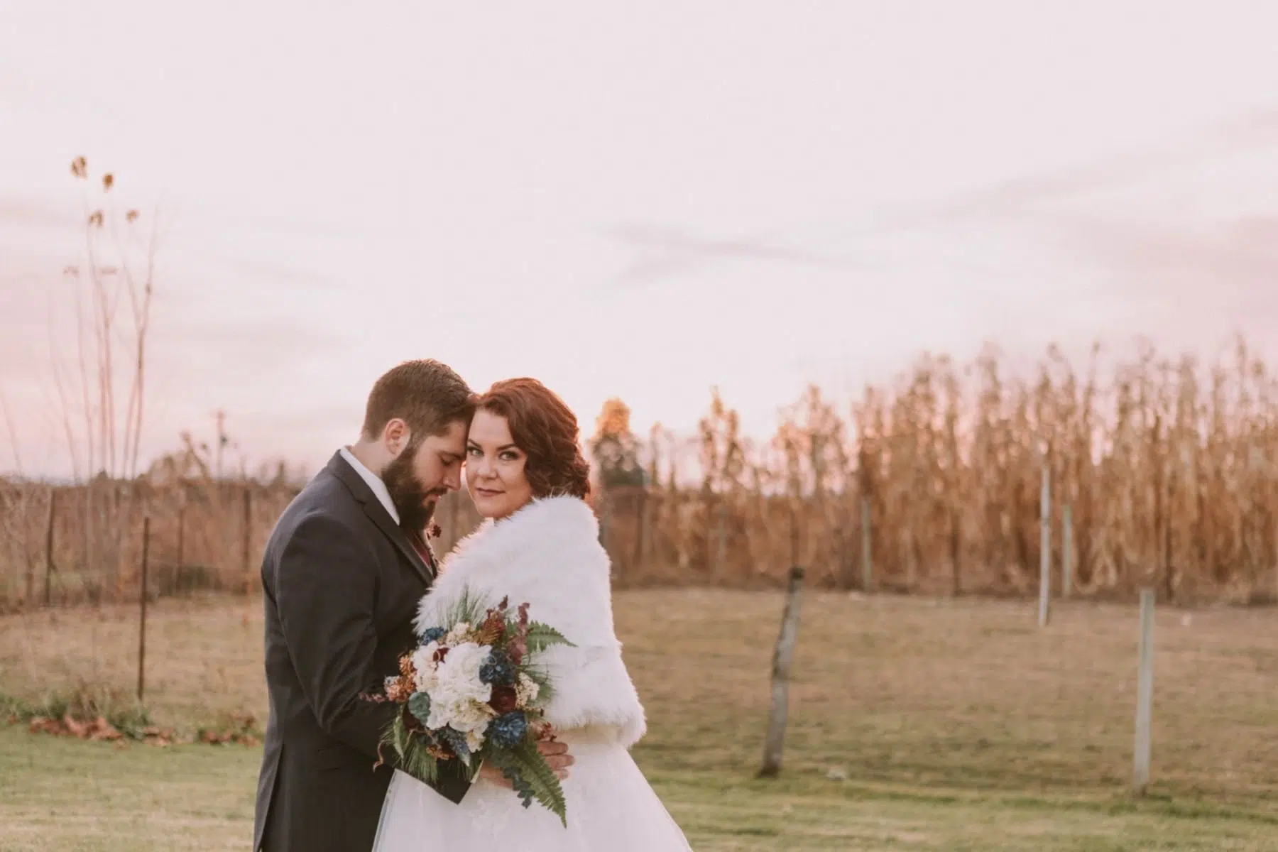 Premier Snohomish & Seattle Wedding Photographer | GSquared Weddings Photography A bride in a white fur shawl holds a bouquet, standing close to the groom in a dark suit. They pose outdoors for their Snohomish wedding, framed by tall, dried plants under a pale sky. 47.9129° N, 122.0982° W | Serving Snohomish, Seattle , Orlando & Beyond