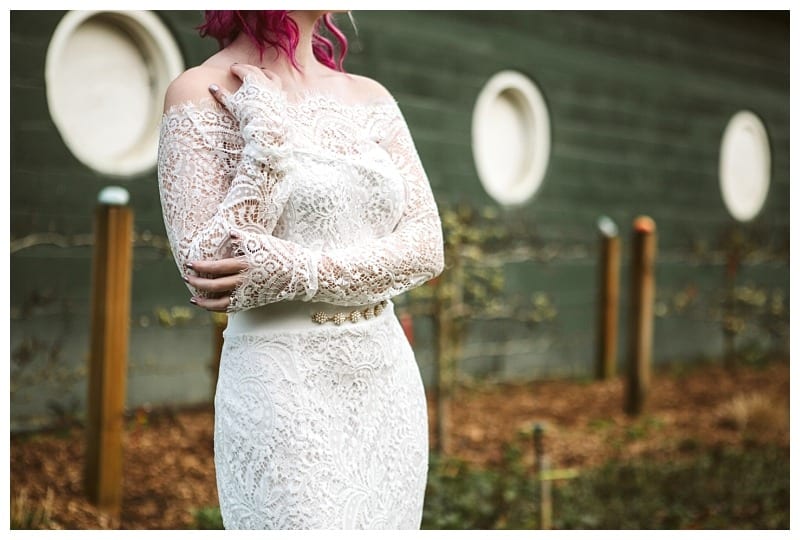Premier Snohomish & Seattle Wedding Photographer | GSquared Weddings Photography A woman with pink hair wears an off-the-shoulder, long-sleeved lace wedding dress, standing outdoors near a green wall with round windows at her Snohomish Seattle wedding, with a garden in the background.                                   | Serving Snohomish, Seattle ,                  & Beyond