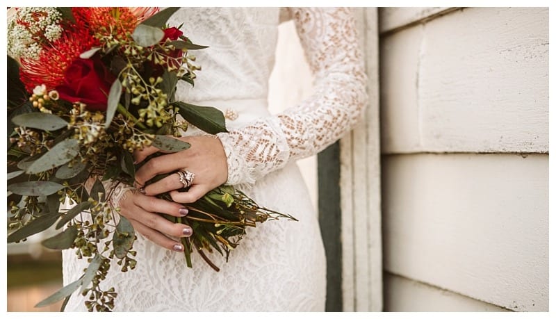 Premier snohomish & seattle wedding photographer | gsquared weddings photography a woman in a long sleeved lace wedding dress holds a bouquet of red and green flowers showing her wedding ring She stands against a white wooden wall capturing a romantic snohomish seattle wedding moment | serving snohomish seattle                   & beyond