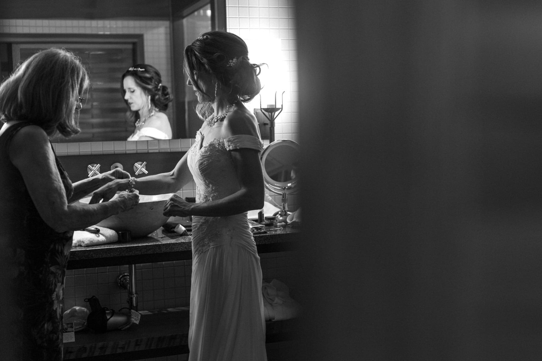 A bride in a white dress stands at a bathroom sink, assisted by an older woman. Their reflections are visible in the mirror. The intimate, softly lit scene unfolds in a Snohomish home, with a blurred foreground framing the moment. Snohomish Wedding Photography