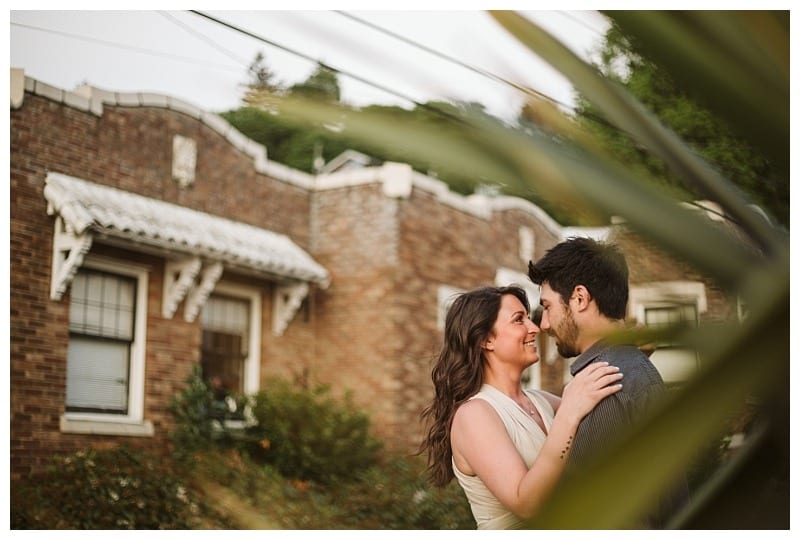 Premier snohomish & seattle wedding photographer | gsquared weddings photography a couple stands close together smiling and gazing into each others eyes in front of a brick house with greenerya perfect snohomish seattle engagement moment Blurred leaves frame the romantic scene | serving snohomish seattle                   & beyond