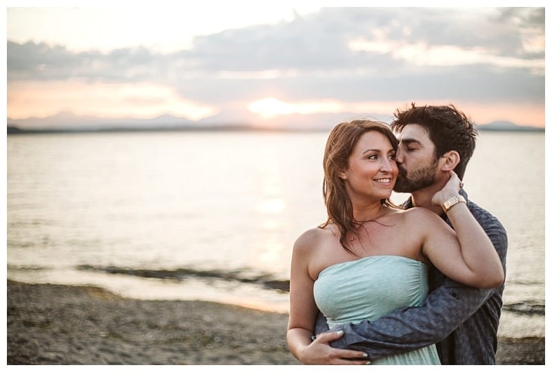 Premier snohomish & seattle wedding photographer | gsquared weddings photography a couple embraces on a beach at sunset during their snohomish seattle engagement The woman smiles while the man stands behind her gently kissing her cheek as the ocean sparkles under a cloudy sky with the sun setting in the background | serving snohomish seattle                   & beyond