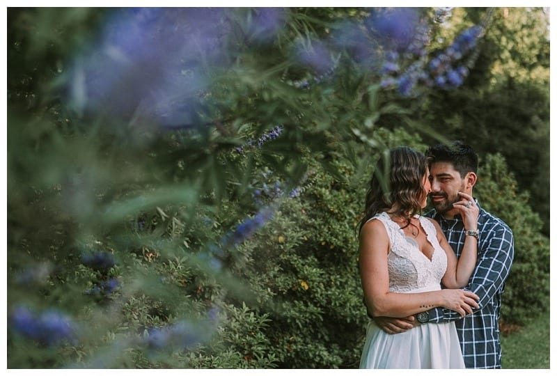 Premier snohomish & seattle wedding photographer | gsquared weddings photography a couple embraces in a garden surrounded by greenery and purple flowers during their snohomish seattle wedding engagement The woman wears a white dress as the man gently touches her face both smiling Soft focus flowers frame the left foreground | serving snohomish seattle                   & beyond
