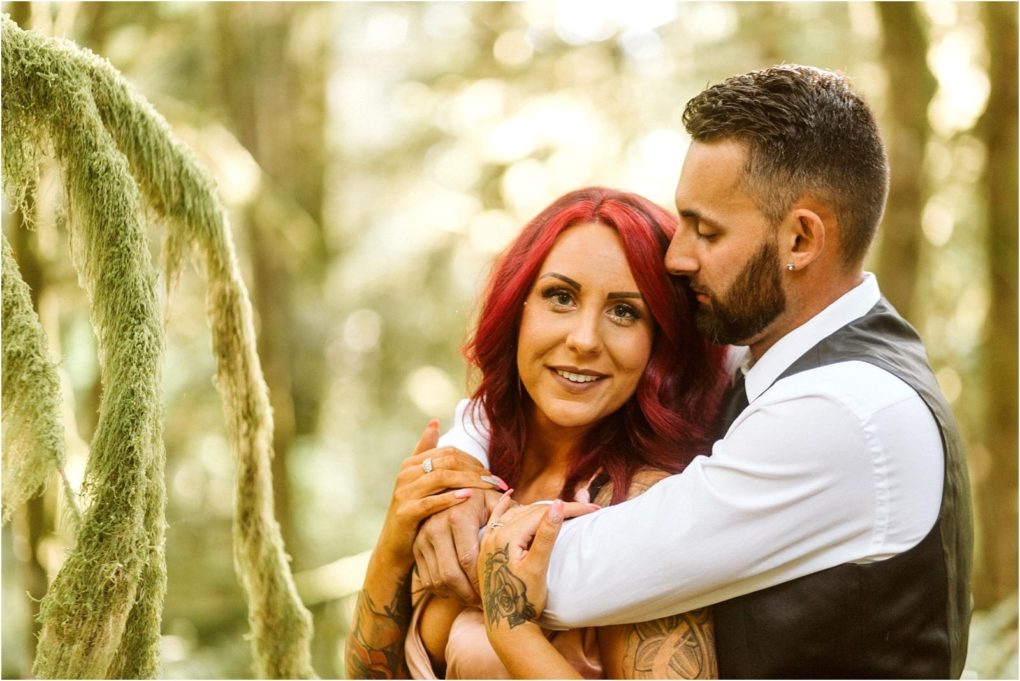 Premier snohomish & seattle wedding photographer | gsquared weddings photography a man with dark hair and a beard hugs a woman with long red hair and tattoos from behind in a sunlit forest near snohomish both looking content The woman smiles at the camera while the man gently rests his head near hers | serving snohomish seattle                   & beyond