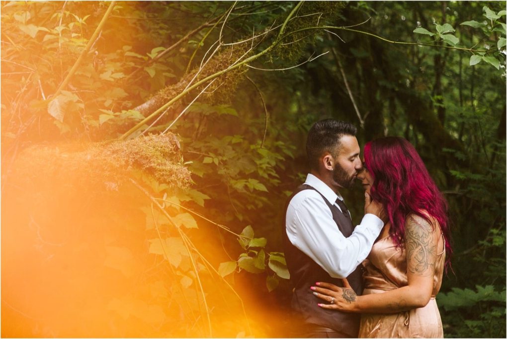Premier snohomish & seattle wedding photographer | gsquared weddings photography a couple stands closely in a lush green forest near snohomish The man wears a white shirt and vest the woman with long bright reddish hair wears a light dress An orange light flare adds a dreamy touch to this seattle engagement moment | serving snohomish seattle                   & beyond