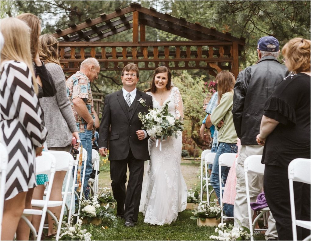 Premier snohomish & seattle wedding photographer | gsquared weddings photography a bride in a white dress and a man in a suit walk arm in arm down an outdoor aisle lined with white chairs and guests at this romantic snohomish wedding with a wooden arbor and lush greenery in the background | serving snohomish seattle                   & beyond