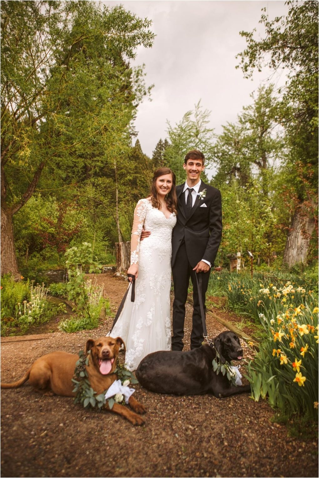 Premier snohomish & seattle wedding photographer | gsquared weddings photography a bride and groom stand smiling in a garden during their snohomish wedding dressed in wedding attire with two large dogs wearing floral collars lying among green plants and yellow flowers | serving snohomish seattle                   & beyond