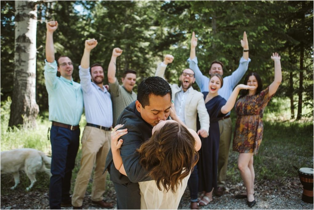 Premier snohomish & seattle wedding photographer | gsquared weddings photography a couple kisses in the foreground while a group of joyful friends cheers with raised arms behind them among treesa sweet snohomish seattle wedding engagement moment A dog is partly visible on the left side of the image | serving snohomish seattle                   & beyond