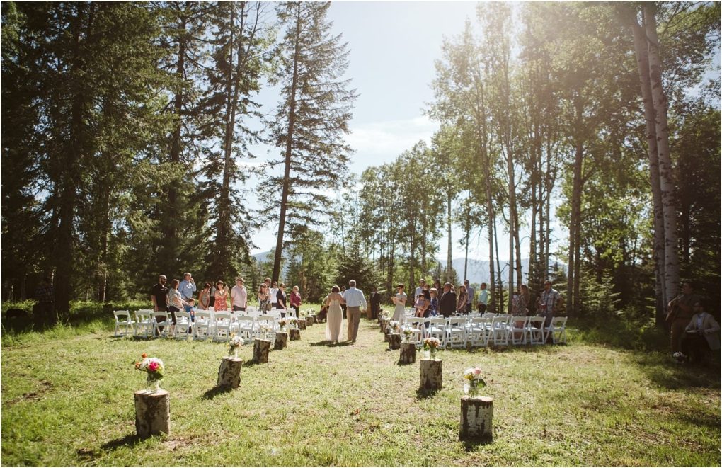 Premier snohomish & seattle wedding photographer | gsquared weddings photography an outdoor wedding ceremony setup in a forest clearing near snohomish with white chairs arranged in rows guests standing or seated and small tree stumps with flower arrangements lining the aisle Sunlight filters through tall trees | serving snohomish seattle                   & beyond