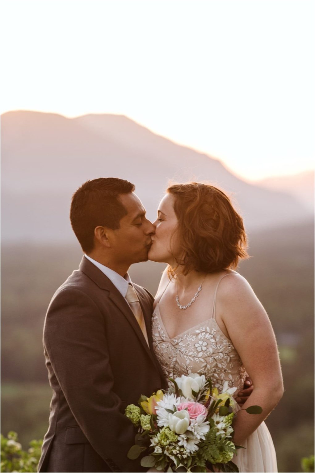 Premier snohomish & seattle wedding photographer | gsquared weddings photography a bride and groom kiss outdoors at sunset during their snohomish wedding with mountains blurred in the background The bride holds a bouquet of flowers and wears a beaded dress while the groom wears a suit and tie | serving snohomish seattle                   & beyond