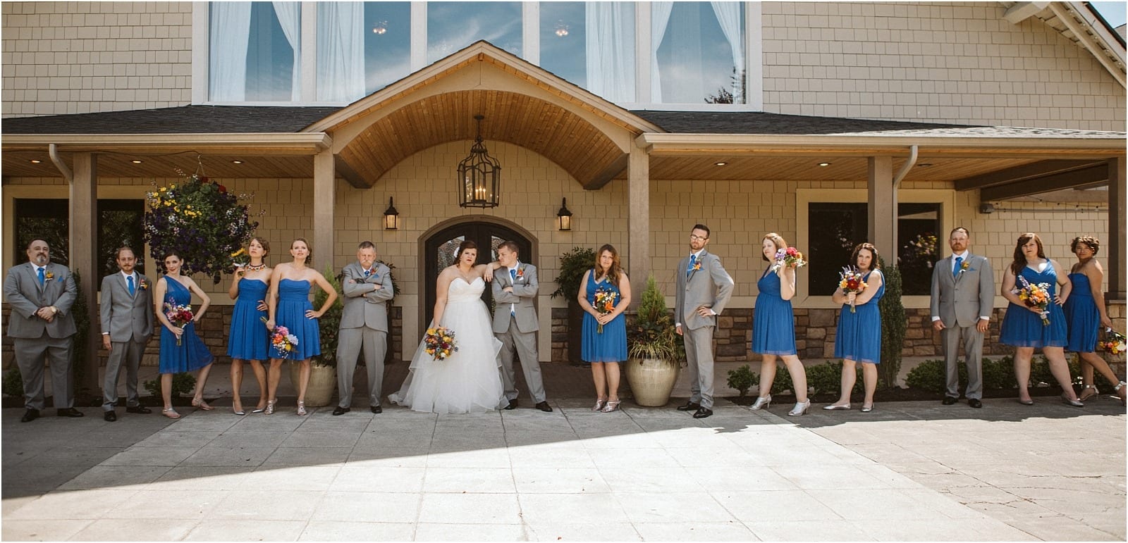 Premier Snohomish & Seattle Wedding Photographer | GSquared Weddings Photography A Snohomish wedding party poses outside a tan building. The bride and groom stand in the center, surrounded by bridesmaids in blue dresses and groomsmen in gray suits, all holding colorful bouquets and smiling.                                   | Serving Snohomish, Seattle ,                  & Beyond