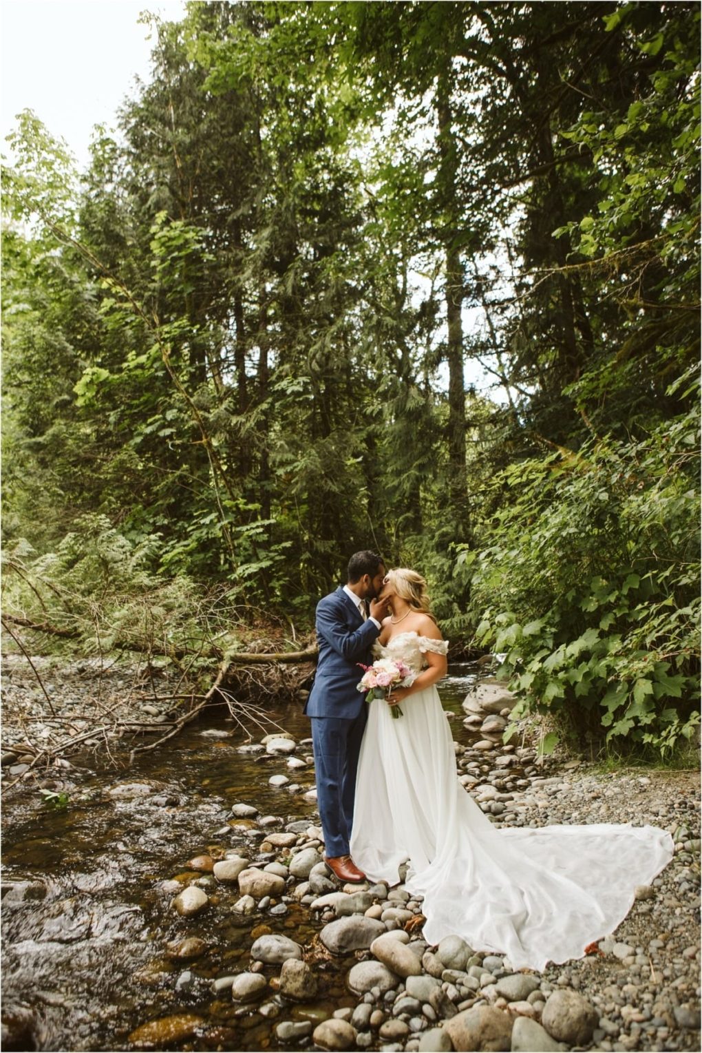 Premier snohomish & seattle wedding photographer | gsquared weddings photography a bride and groom stand by a rocky stream in a lush green forest for their snohomish wedding The groom in a blue suit kisses the bride who wears a white gown and holds a bouquet as her train trails behind her on the stones | serving snohomish seattle                   & beyond