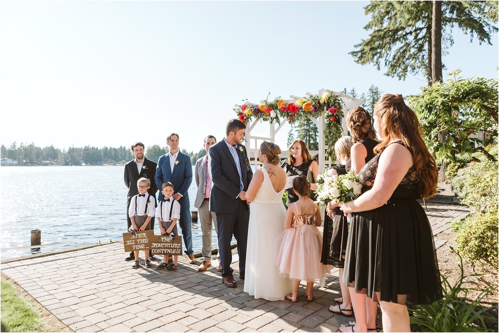 Premier Snohomish & Seattle Wedding Photographer | GSquared Weddings Photography A bride and groom stand under a flower-adorned arch by a lake, holding hands during their sunny Snohomish wedding ceremony. Family and friends watch as two children hold signs, celebrating this beautiful Seattle engagement.                                   | Serving Snohomish, Seattle ,                  & Beyond