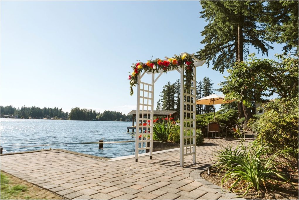 Premier snohomish & seattle wedding photographer | gsquared weddings photography a white lattice wedding arch decorated with colorful flowers stands on a stone patio by a peaceful lake in snohomish surrounded by greenery and tall treesan enchanting setting for your seattle wedding or engagement | serving snohomish seattle                   & beyond