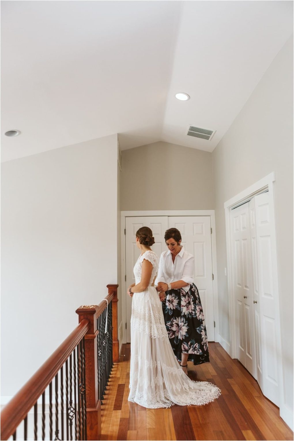Premier snohomish & seattle wedding photographer | gsquared weddings photography a woman in a white bridal gown stands by a staircase as another woman dressed in a white blouse and floral skirt helps fasten her dress in a bright hallway with wooden floorscapturing a warm snohomish wedding moment near seattle | serving snohomish seattle                   & beyond