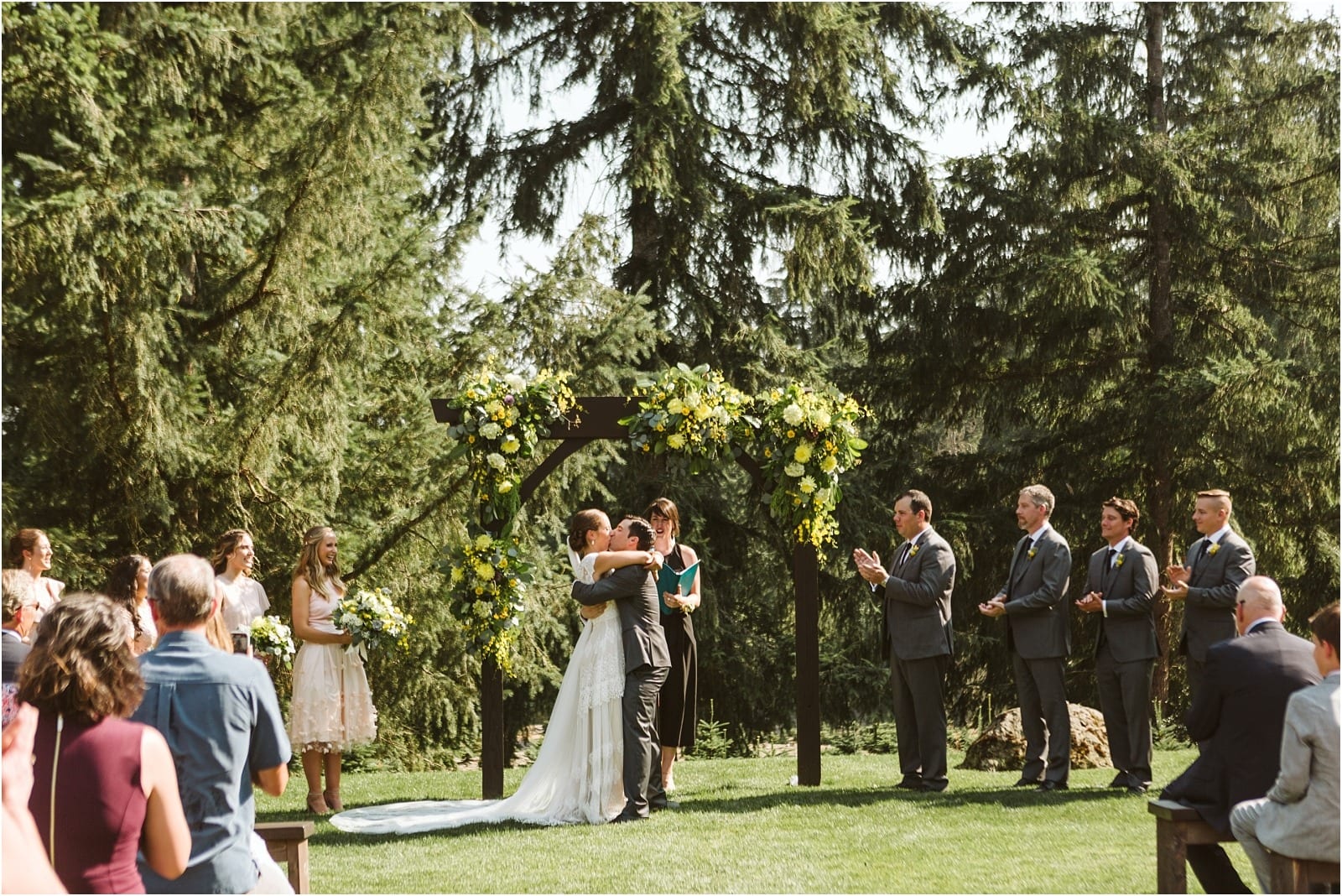 Premier Snohomish & Seattle Wedding Photographer | GSquared Weddings Photography A bride and groom embrace under a floral arch during a Snohomish outdoor wedding ceremony surrounded by trees, while their wedding party and guests watch and applaud.                                   | Serving Snohomish, Seattle ,                  & Beyond