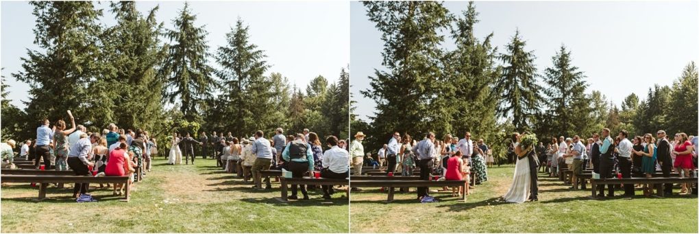 Premier snohomish & seattle wedding photographer | gsquared weddings photography wedding guests stand and clap outdoors on a sunny day in snohomish gathered beside a grassy aisle lined with large pine trees A joyful bride and groom walk down the aisle celebrating their wedding after the ceremony | serving snohomish seattle                   & beyond