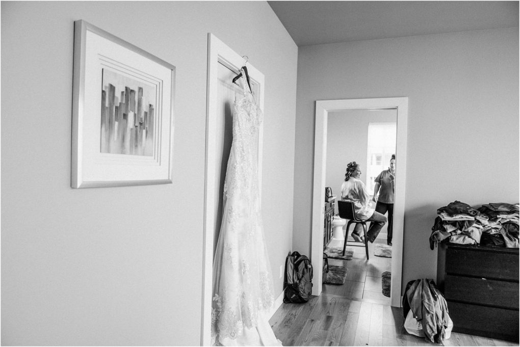 Premier snohomish & seattle wedding photographer | gsquared weddings photography a lacy wedding dress hangs on a door in a tidy room near snohomish Through an open doorway two people sit facing each other one in hair rollers preparing for a seattle wedding The scene is calm and full of anticipation | serving snohomish seattle                   & beyond
