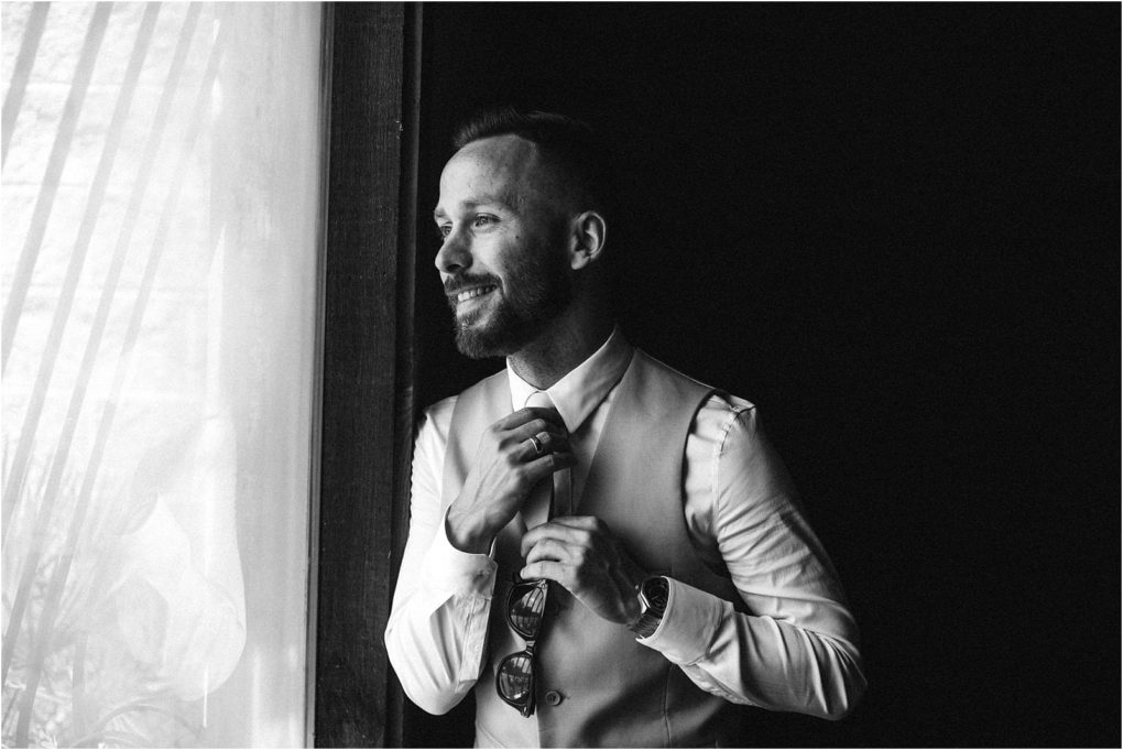 Premier snohomish & seattle wedding photographer | gsquared weddings photography black and white photo of a man smiling as he looks out a window He is wearing a suit vest and adjusting his tie with soft light illuminating his facecapturing a timeless moment from a seattle wedding engagement | serving snohomish seattle                   & beyond