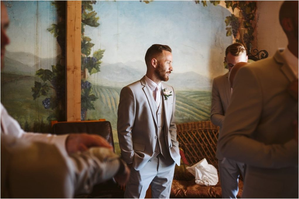Premier snohomish & seattle wedding photographer | gsquared weddings photography a man in a light gray suit stands with hands in his pockets smiling in a rustic room near snohomish with a mural of fields and mountainsperfect for a seattle wedding or engagement Other men in similar suits are partially visible around him | serving snohomish seattle                   & beyond